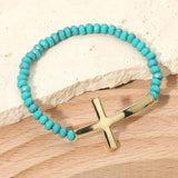 Turquoise beaded bracelet with a gold cross charm on a wooden surface.