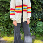 Person wearing a colorful striped cardigan and dark pants standing outdoors with greenery in the background
