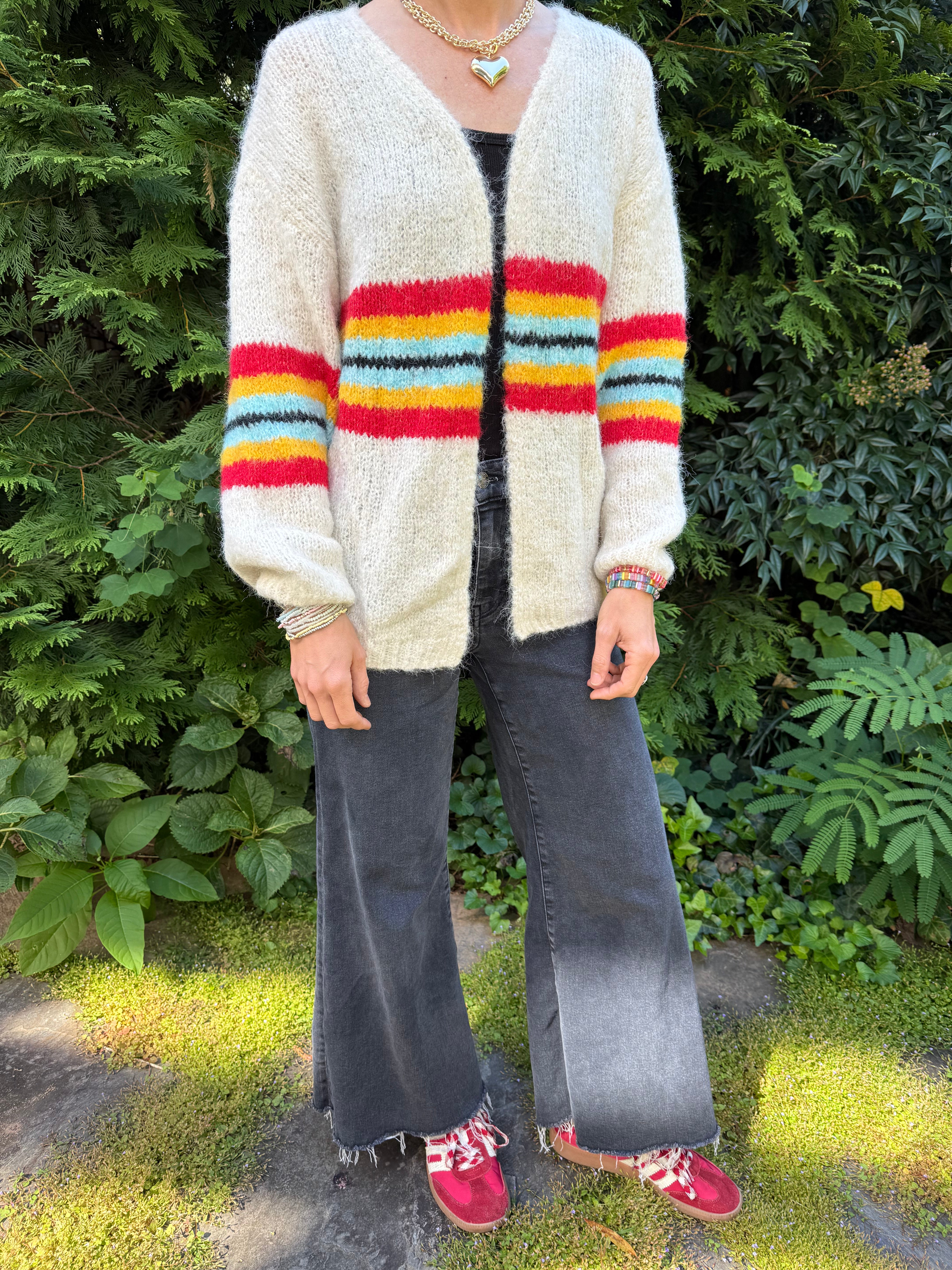 Person wearing a colorful striped cardigan and dark pants standing outdoors with greenery in the background