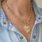 Gold necklace with a large heart pendant worn by a person in a light blue denim shirt.