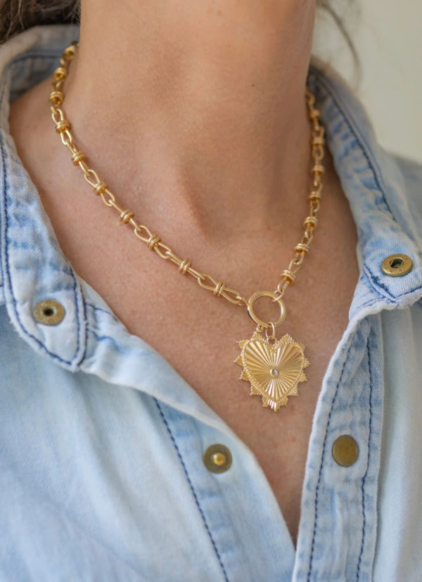 Gold necklace with a large heart pendant worn by a person in a light blue denim shirt.